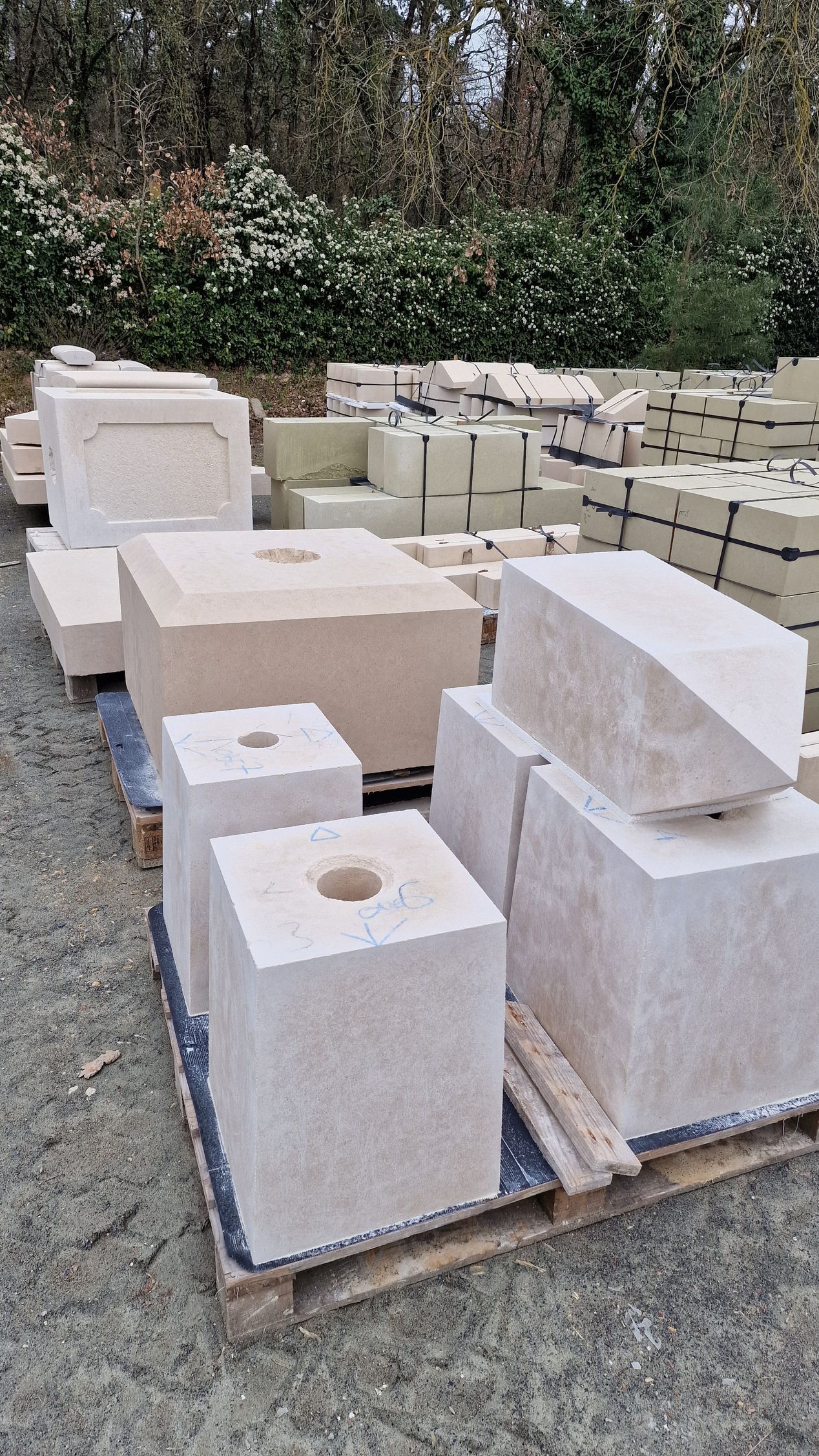 Une palette de blocs et de piliers en pierre claire sculptée est installée à l'extérieur, sur un terrain gravillonné avec des arbres en arrière-plan.