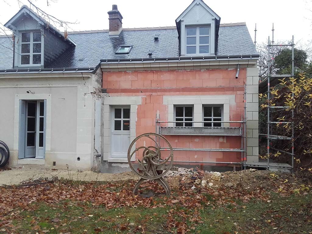 Une maison à deux étages avec une extension en briques rouges partiellement rénovée, une porte blanche, des échafaudages et des feuilles d'automne tombées.