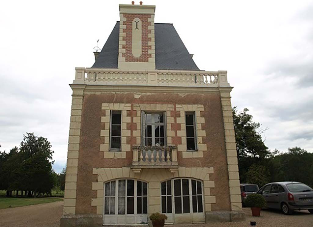 Un bâtiment en pierre de deux étages avec un toit en ardoise, doté d'un balcon, de fenêtres cintrées et d'une voiture garée sur la droite.