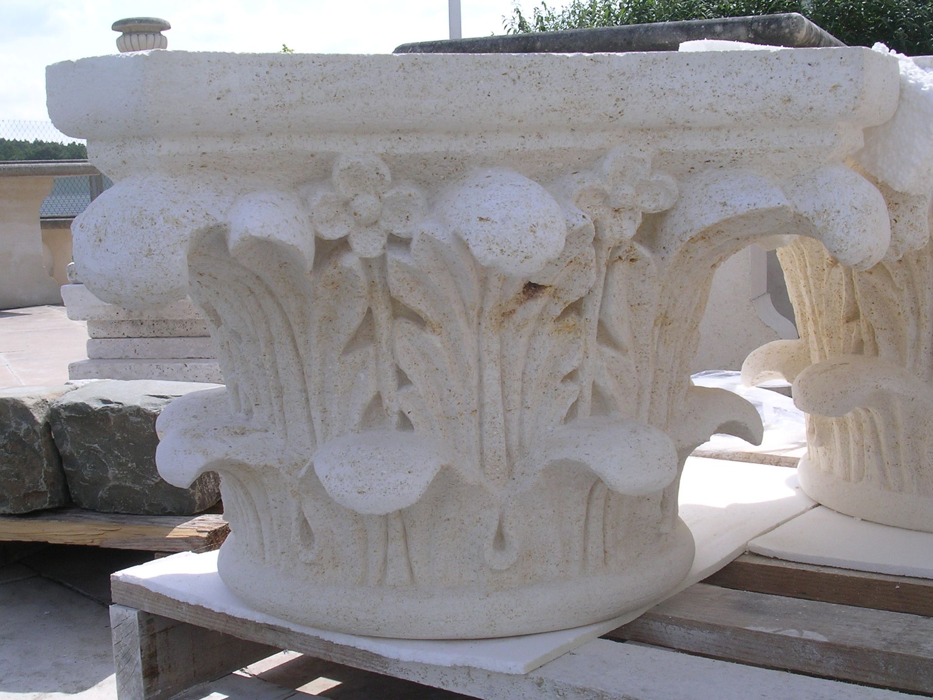 Un chapiteau de colonne en pierre claire sculpté de motifs floraux et de feuilles, posé sur une palette en bois à l'extérieur.