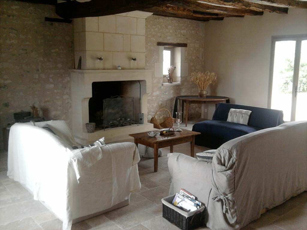 Un salon rustique et chaleureux avec des murs en pierre, une grande cheminée, des poutres apparentes au plafond et trois canapés autour d'une table basse.