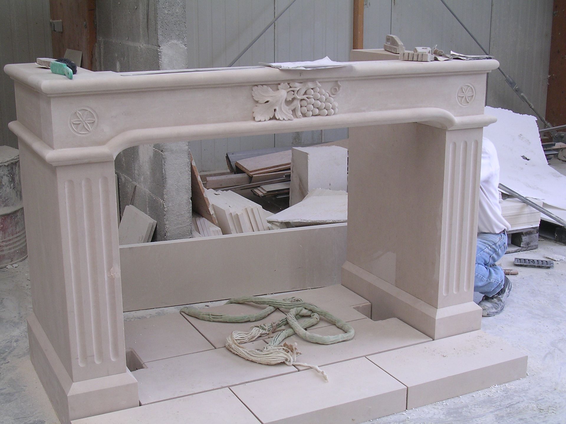 Dans un atelier, une cheminée en pierre claire partiellement assemblée, ornée de détails floraux sculptés, se trouve à proximité.