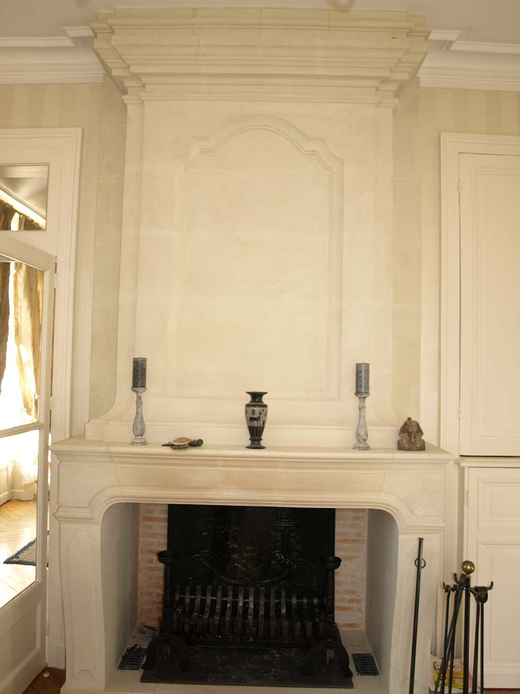 Une cheminée en pierre calcaire blanche, ornée d'un manteau, d'un foyer et d'une grille en fer finement travaillés, est encadrée par des objets décoratifs dans une pièce.