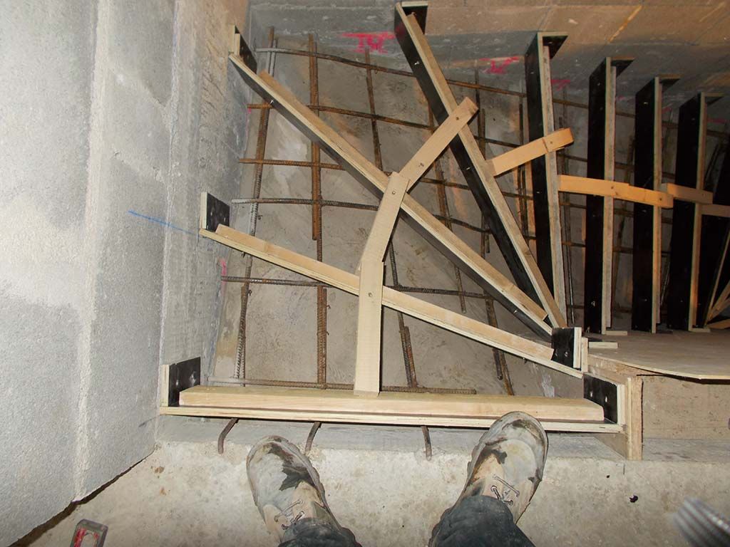 Vue subjective de bottes de chantier devant une structure de coffrage en bois destinée à recevoir un escalier en béton.