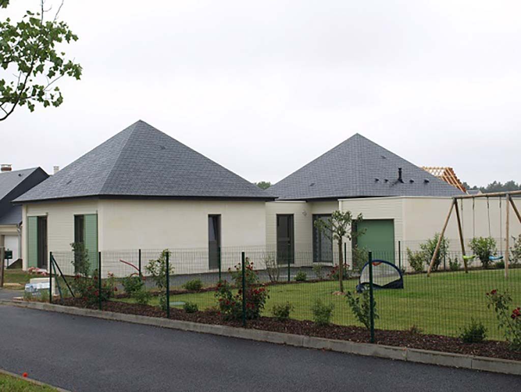 Deux maisons de plain-pied aux toits à quatre pans gris foncé et aux murs couleur crème, séparées par une pelouse verte et une clôture en fil de fer.