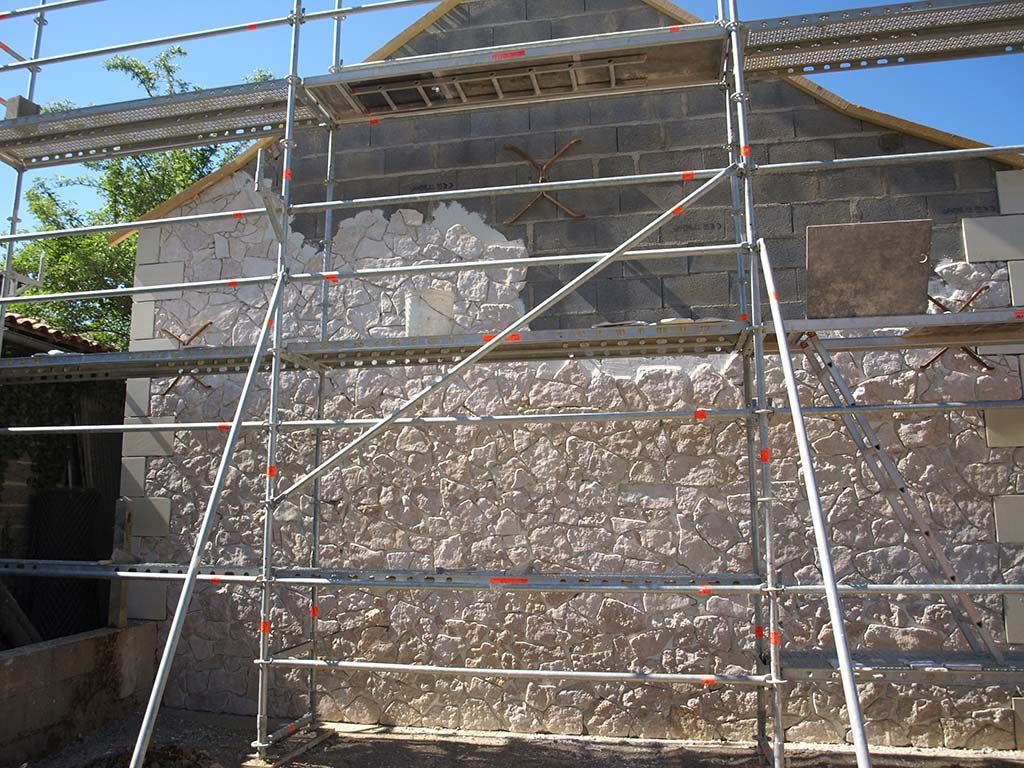 Sous un ciel bleu limpide, une structure d'échafaudage métallique se dresse contre un mur de pierre partiellement enduit.