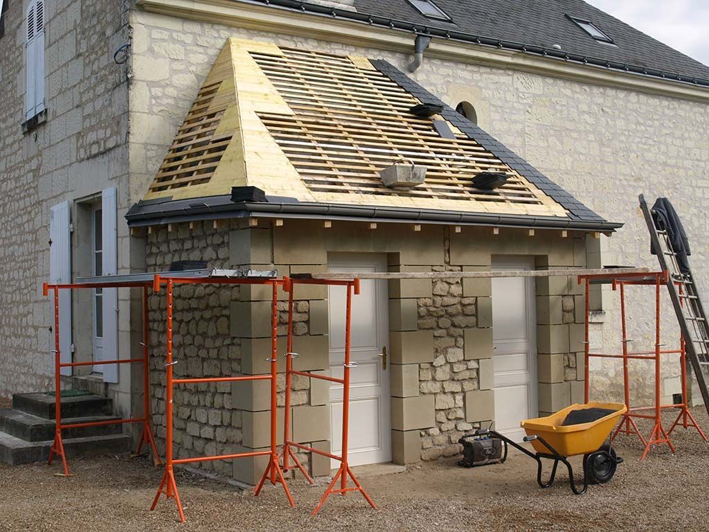 Une structure en pierre en cours de rénovation, avec une charpente de toit en bois partiellement apparente, des échafaudages orange et une brouette jaune.