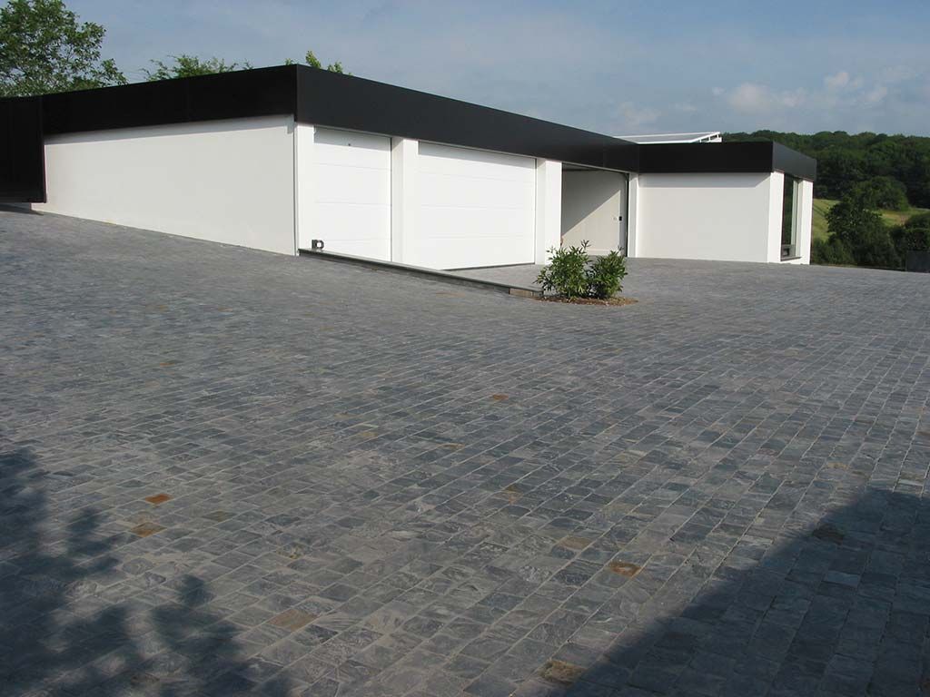 Bâtiment moderne aux murs blancs et au toit noir, adossé à une grande allée pavée de pierre grise.
