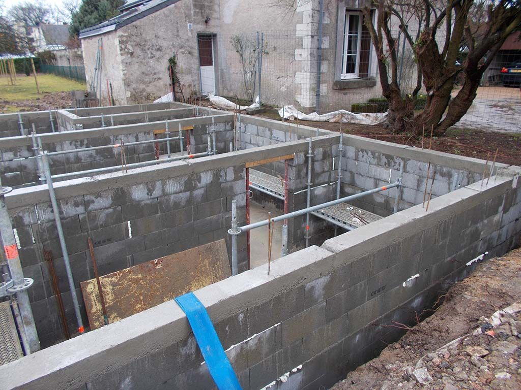 Chantier de construction montrant des murs en blocs de béton gris en cours d'édification à l'aide d'échafaudages métalliques dans une cour arrière résidentielle.