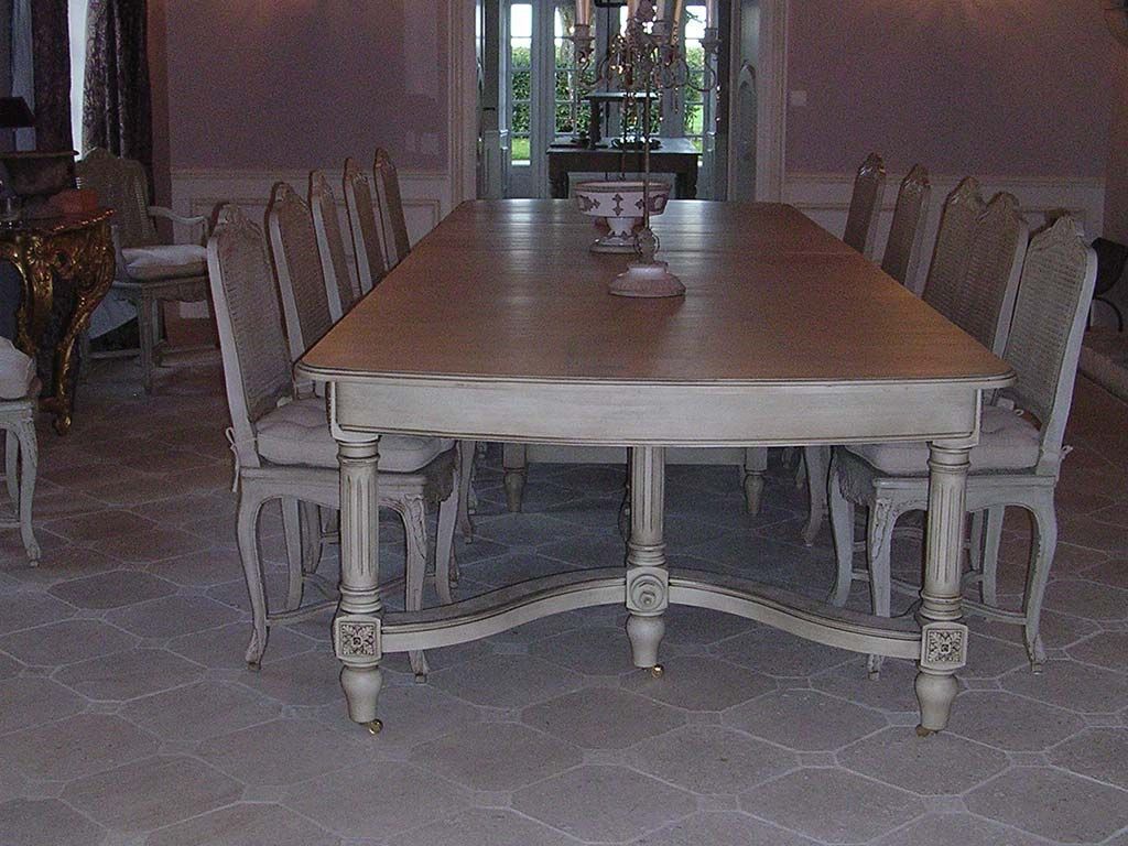 Une longue table à manger en bois gris clair vieilli, avec des chaises assorties, dans une salle à manger élégante et baignée de lumière.