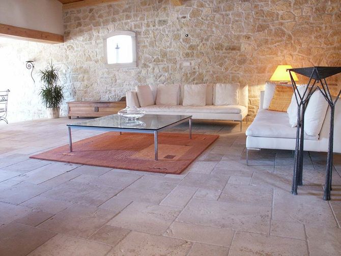 Un salon avec des murs en pierre, des canapés clairs, une table basse en verre et un tapis orange sur un sol carrelé en pierre.