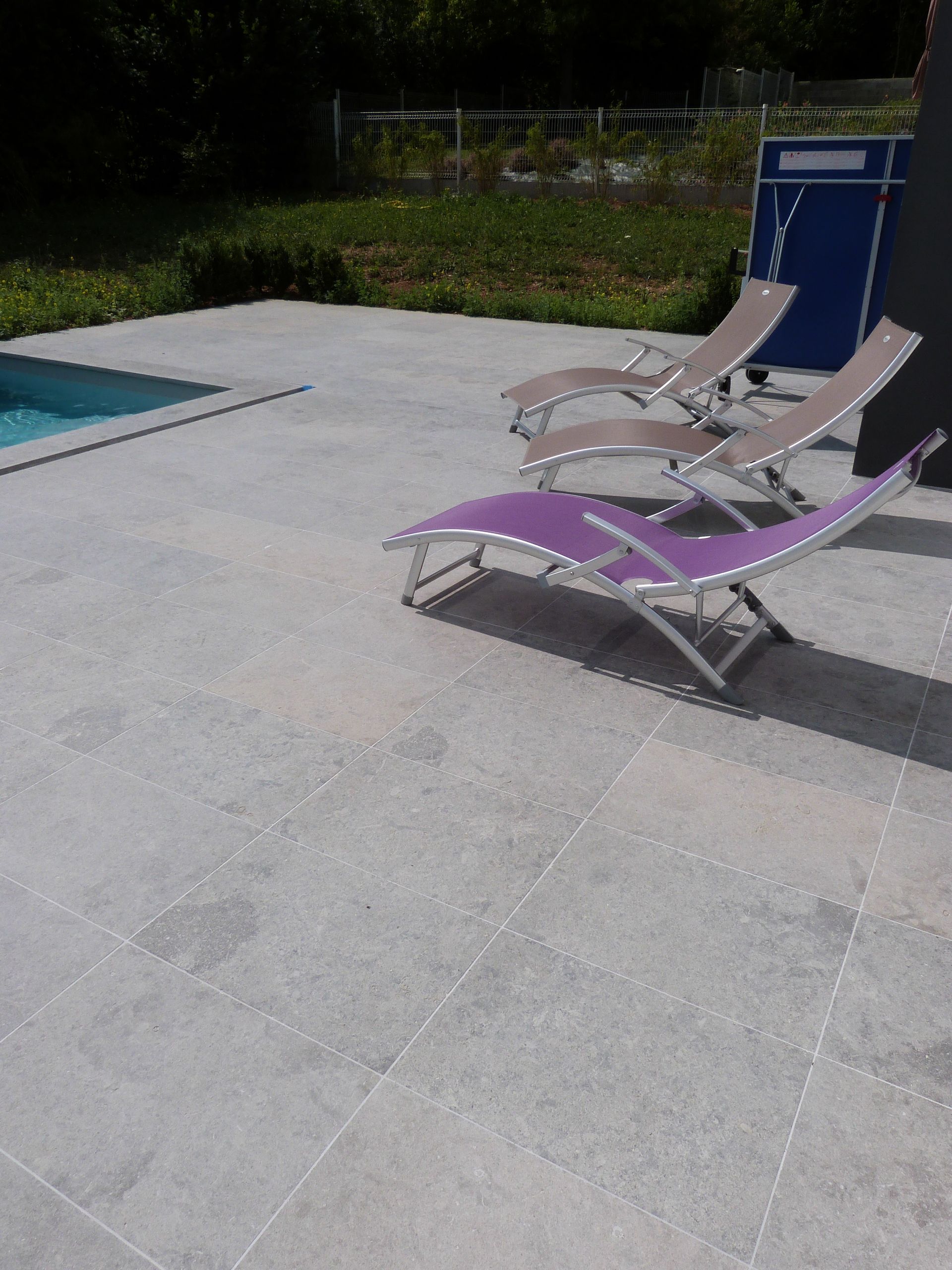 Trois chaises longues — deux beiges et une violette — sont installées sur une terrasse en pierre gris clair, à côté d'une piscine.