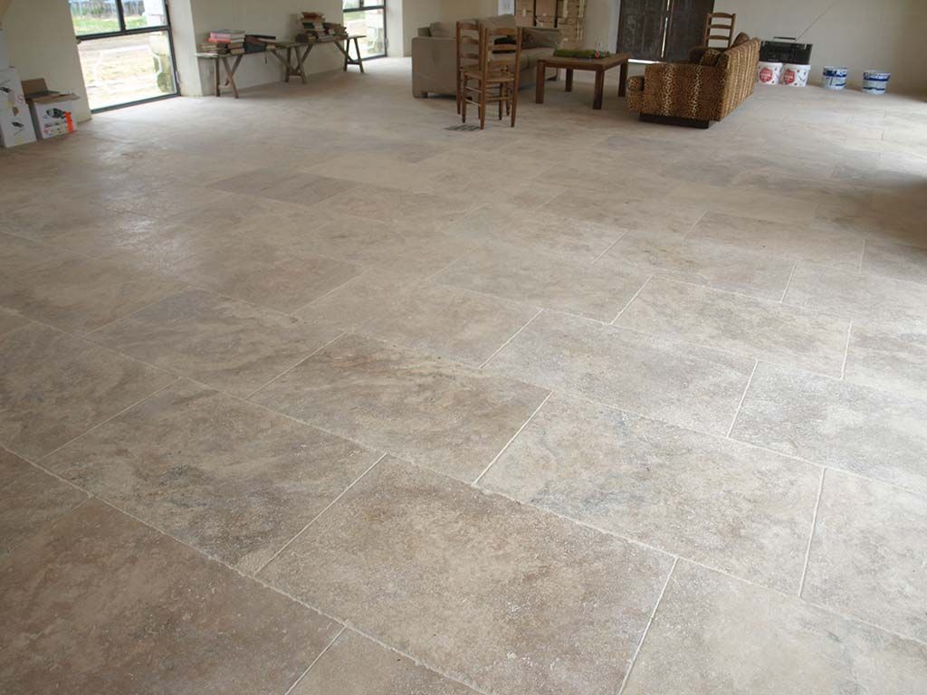 Une pièce spacieuse présente un sol carrelé de grands carreaux de pierre beiges disposés de façon aléatoire, avec des meubles au fond.