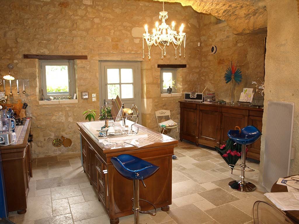 Une pièce aux murs de pierre avec un lustre, une grande table en bois et deux tabourets de bar bleus sur un sol carrelé.