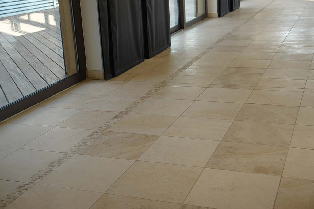 Des carreaux de sol beiges, reliés par une bande de mortier visible et irrégulière, longent un couloir intérieur doté de portes coulissantes en verre.