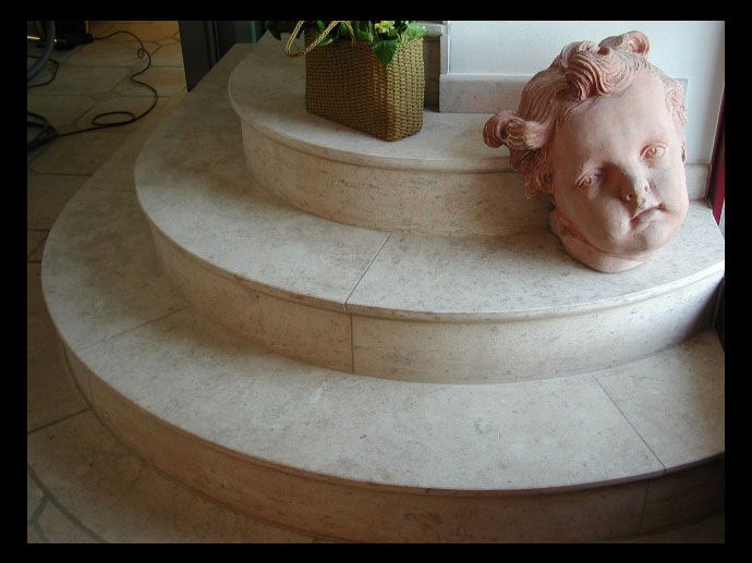 Une sculpture en pierre représentant une tête de chérubin reposant sur un escalier en marbre clair à trois niveaux et à forme incurvée, à côté d'une plante.