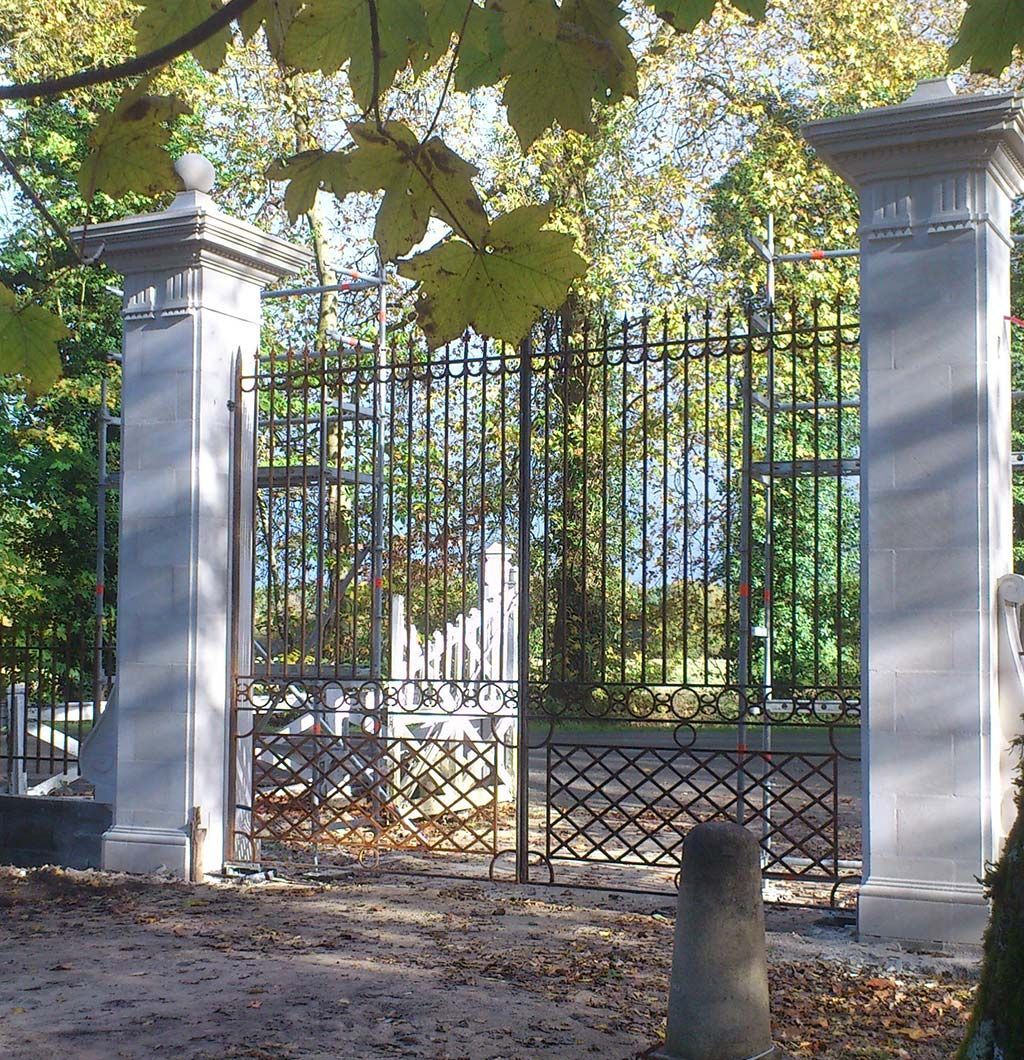 Une grille en fer forgé ouvragée se dresse entre deux hauts piliers de pierre claire, dans un parc tapissé de feuilles d'automne.