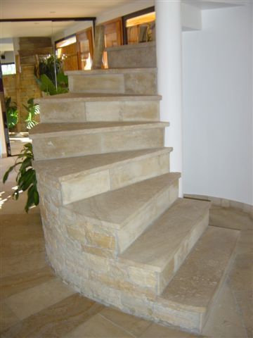 Un escalier courbe construit en blocs de pierre naturelle beige clair, situé dans un intérieur avec un sol carrelé.