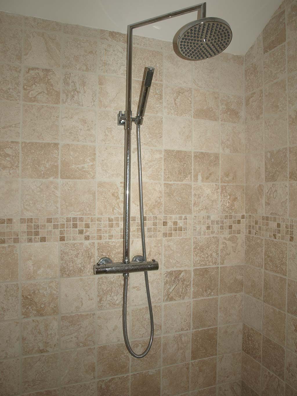 Système de douche à double pommeau chromé, monté sur un mur carrelé de carrelage beige texturé avec une bordure décorative en mosaïque.