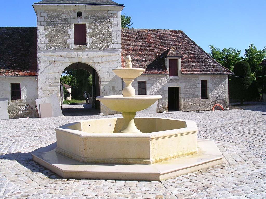 Une fontaine en pierre à plusieurs niveaux se dresse dans une cour pavée, devant un bâtiment historique en pierre doté d'un grand portail voûté.