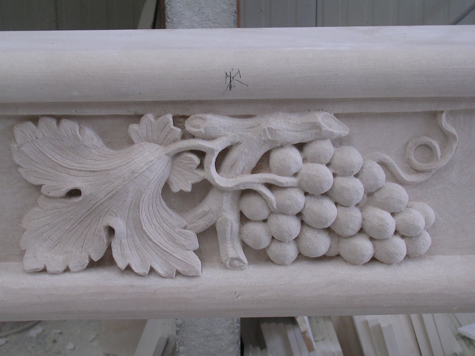 Sculpture en pierre finement travaillée représentant une grappe de raisin avec une grande feuille de vigne sur un rebord architectural de couleur neutre.