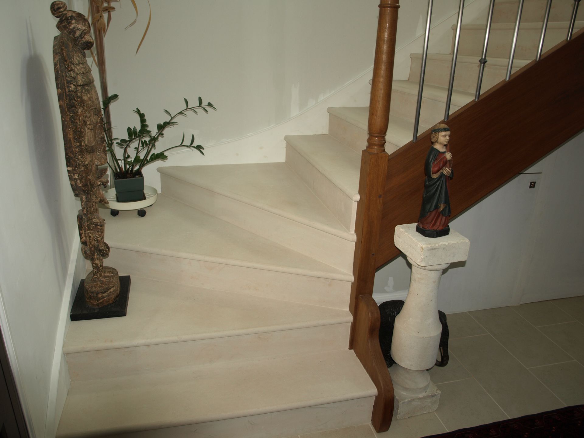 Un escalier en bois aux marches en pierre beige, orné d'une statue sur un piédestal et d'une grande figure décorative contre le mur.