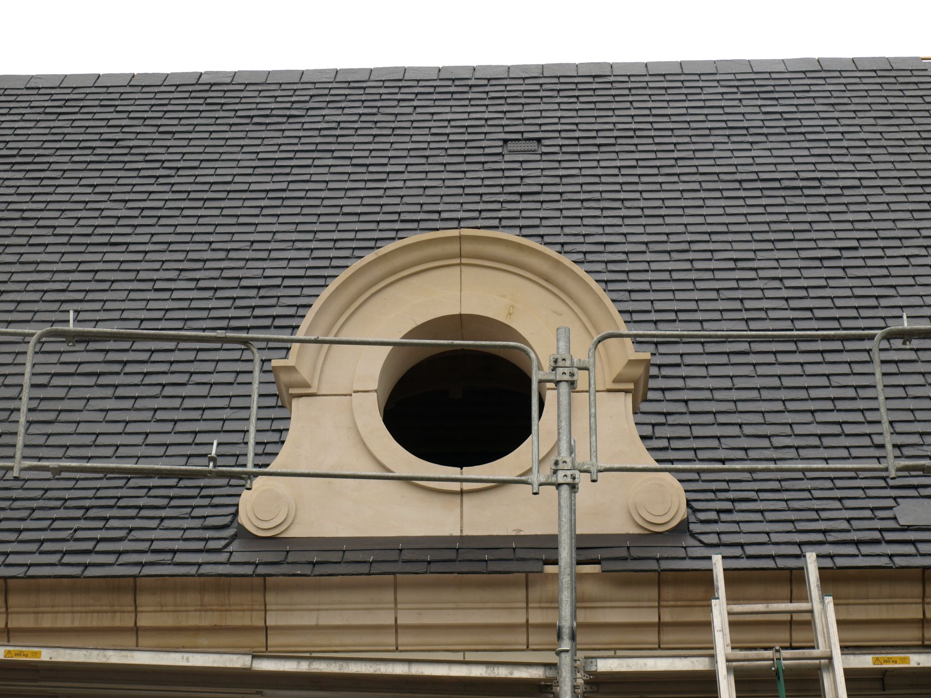 Une fenêtre en oculus, encadrée de pierre décorative, est percée dans un toit d'ardoise gris foncé, visible à travers un échafaudage métallique.