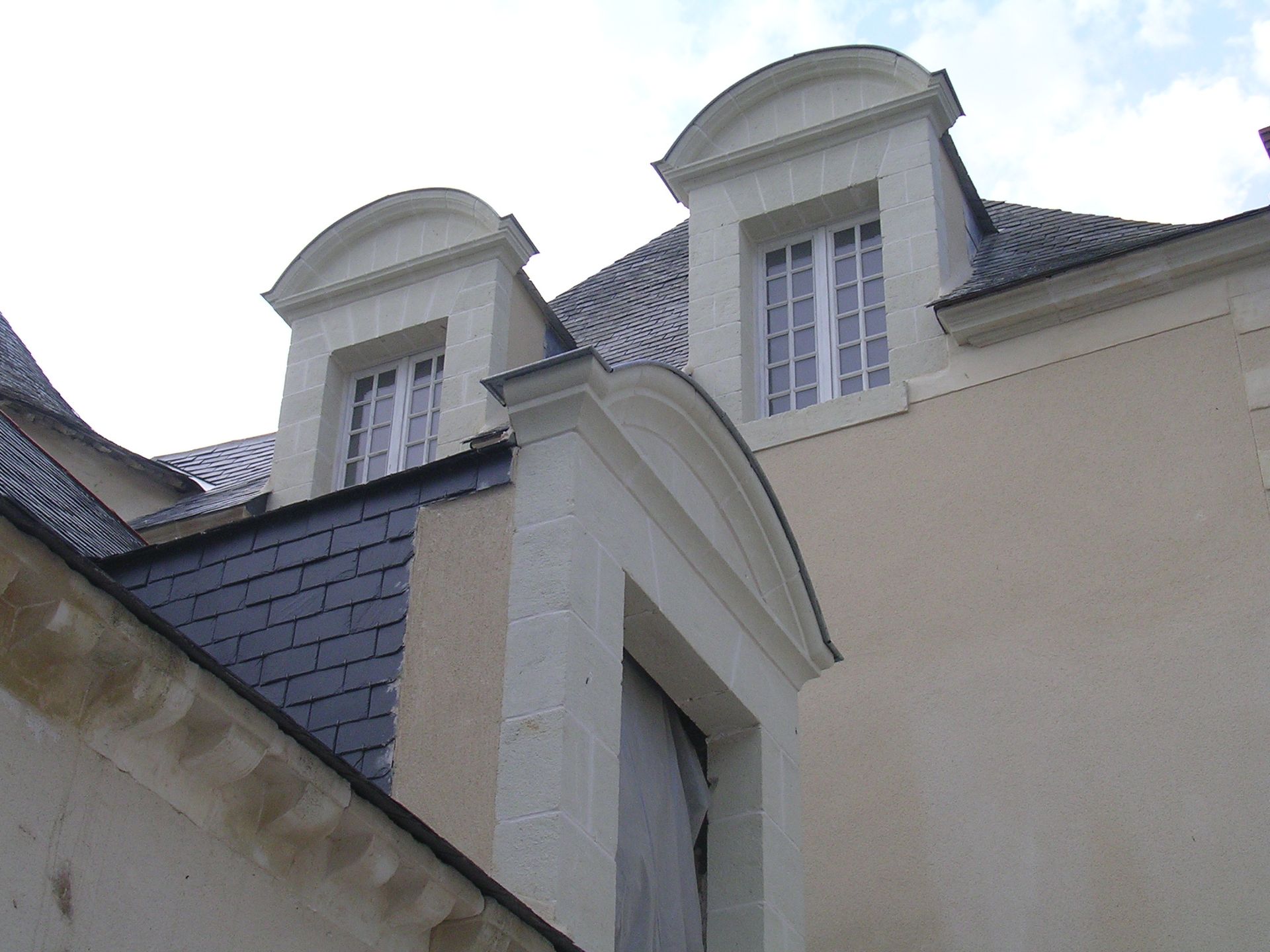 Deux lucarnes en pierre à frontons arqués, encastrées dans un toit d'ardoise, vues d'en bas sur un ciel pâle.