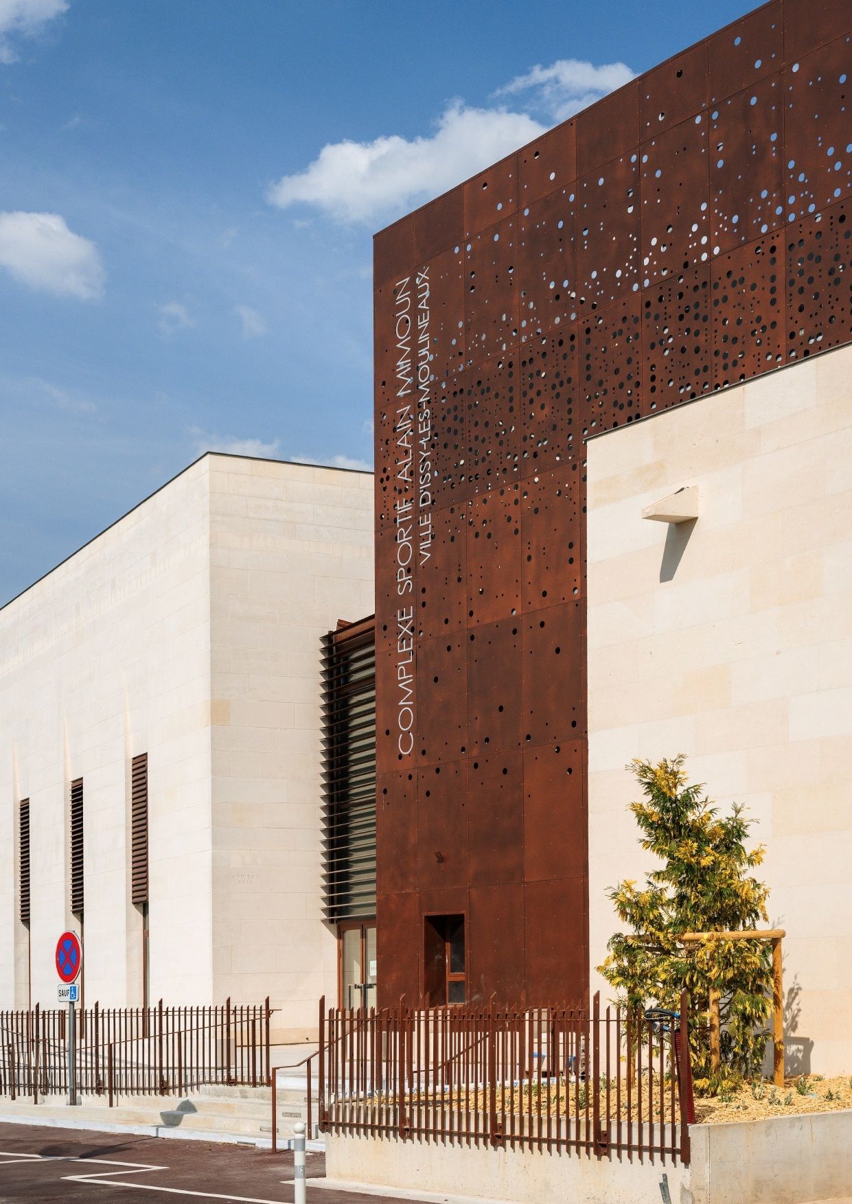 Extérieur de bâtiment moderne avec des murs en calcaire beige contrastant avec une façade métallique verticale perforée de couleur rouille.