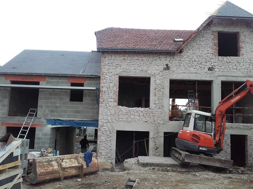 Une pelleteuse orange se dresse devant un chantier de construction où se trouve une maison en pierre et en parpaings de deux étages, partiellement construite.