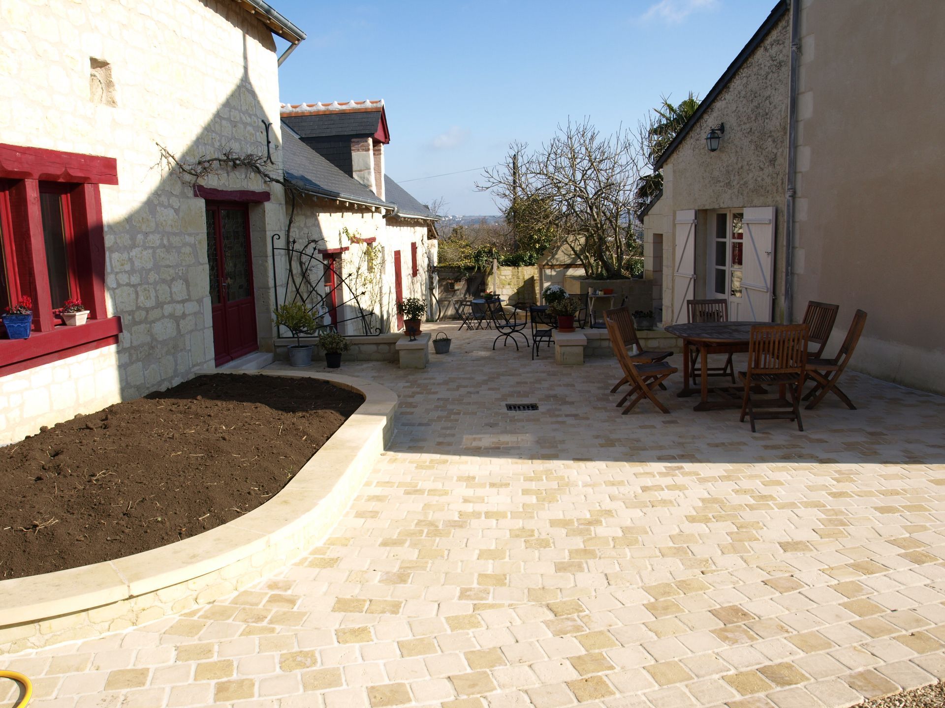 Une cour pavée entre deux bâtiments rustiques, avec une table de patio et des chaises ainsi qu'un parterre de jardin surélevé.