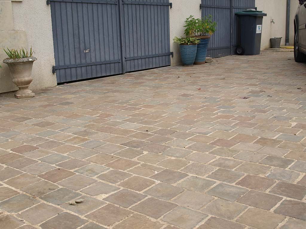 Une allée pavée avec des portes de garage doubles grises, deux plantes en pot et une poubelle sombre contre un mur clair.