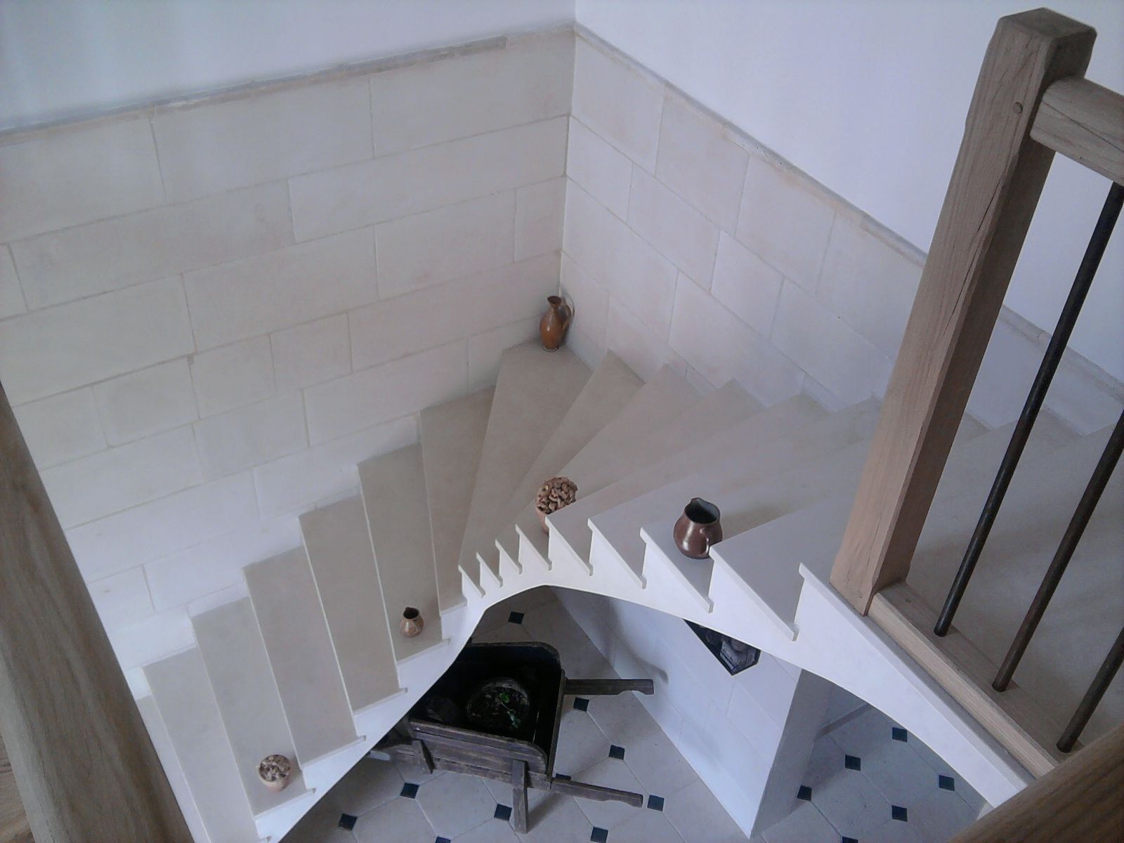 Un escalier courbe de couleur claire, orné d'éléments décoratifs en métal sur chaque marche, descend vers un sol carrelé où se trouve un chariot.