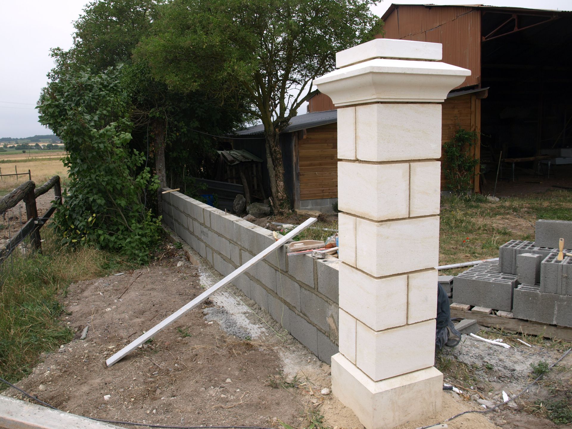 Un pilier en pierre beige se dresse à l'extrémité d'un mur en parpaings de béton gris en construction dans une cour herbeuse.