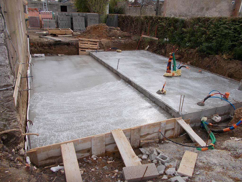 Un chantier de construction montrant deux fondations en béton fraîchement coulées, entourées de terre et de coffrages en bois.