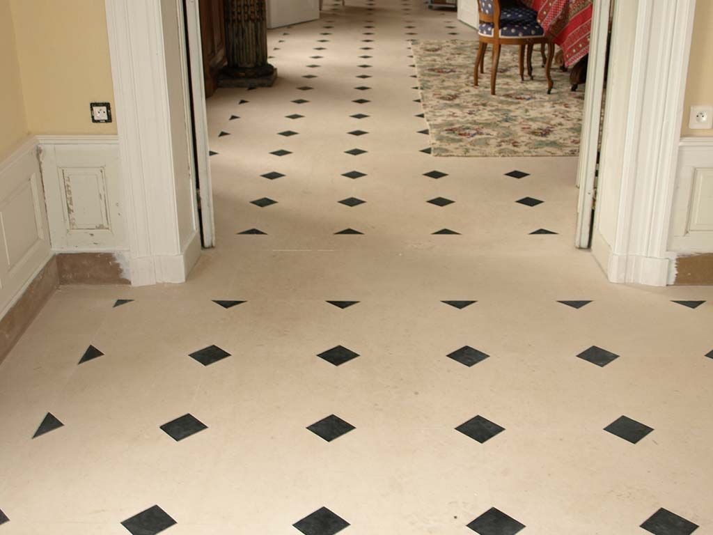 Un sol de couloir recouvert de carreaux carrés beige clair avec des accents noirs plus petits, en forme de losange, régulièrement espacés.