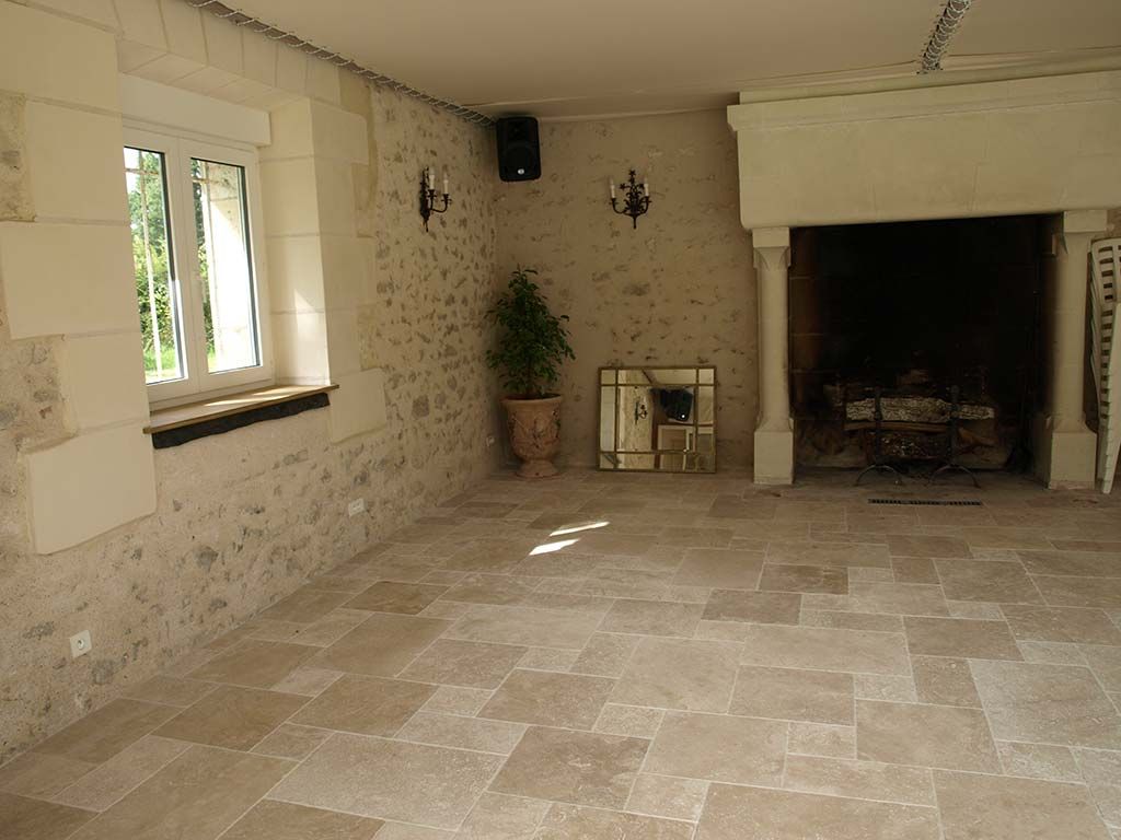 Une pièce aux murs de pierre, au sol carrelé clair, avec une grande cheminée en pierre et une fenêtre ornée d'une plante en pot.