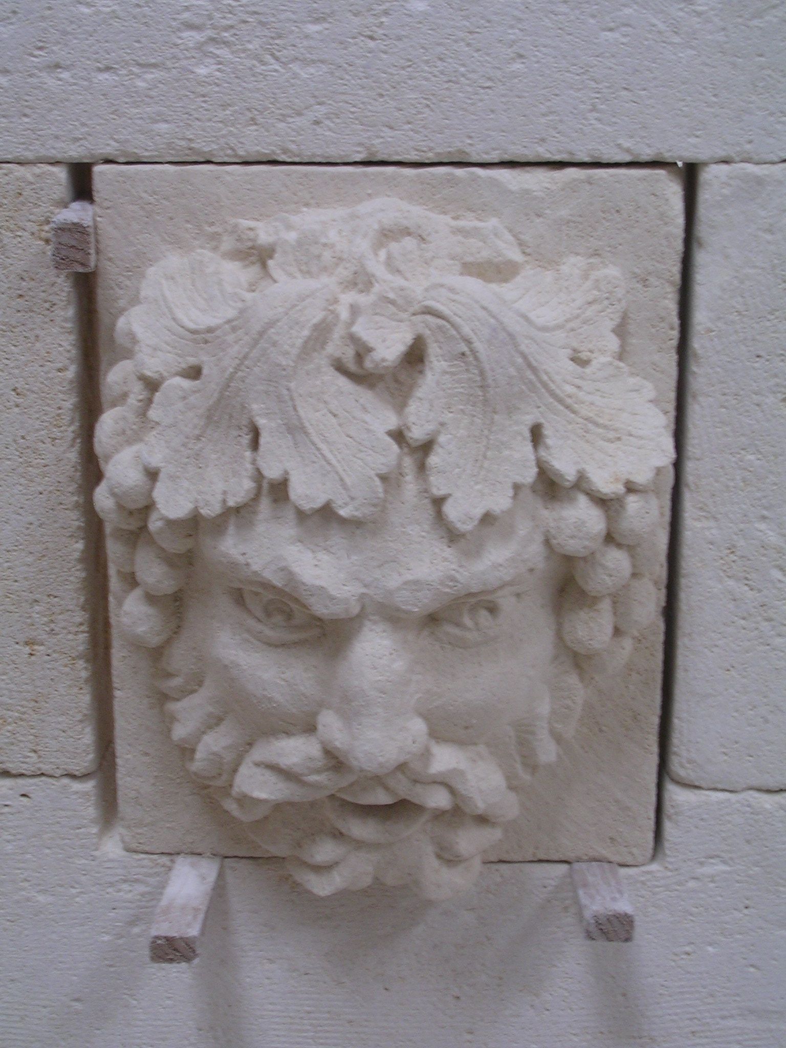 Un bas-relief en pierre représentant un visage barbu orné de feuilles et de grappes de vigne, incrusté dans un mur de maçonnerie claire.