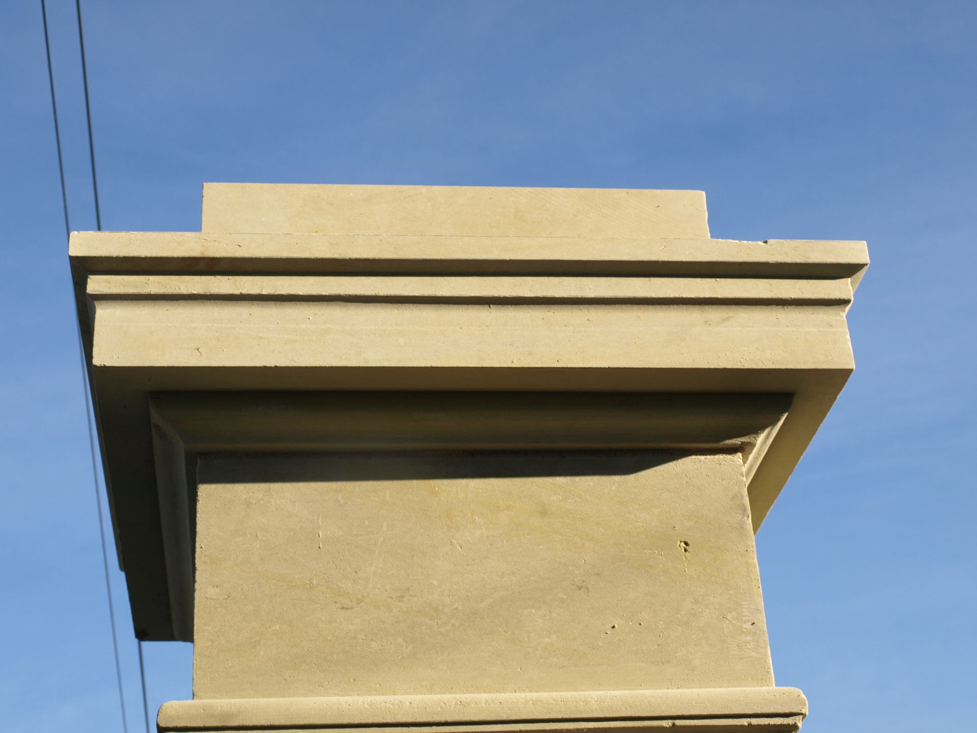 Gros plan sur un chapiteau de pilier en béton moulé beige, aux bords architecturaux étagés, se détachant sur un ciel bleu clair.
