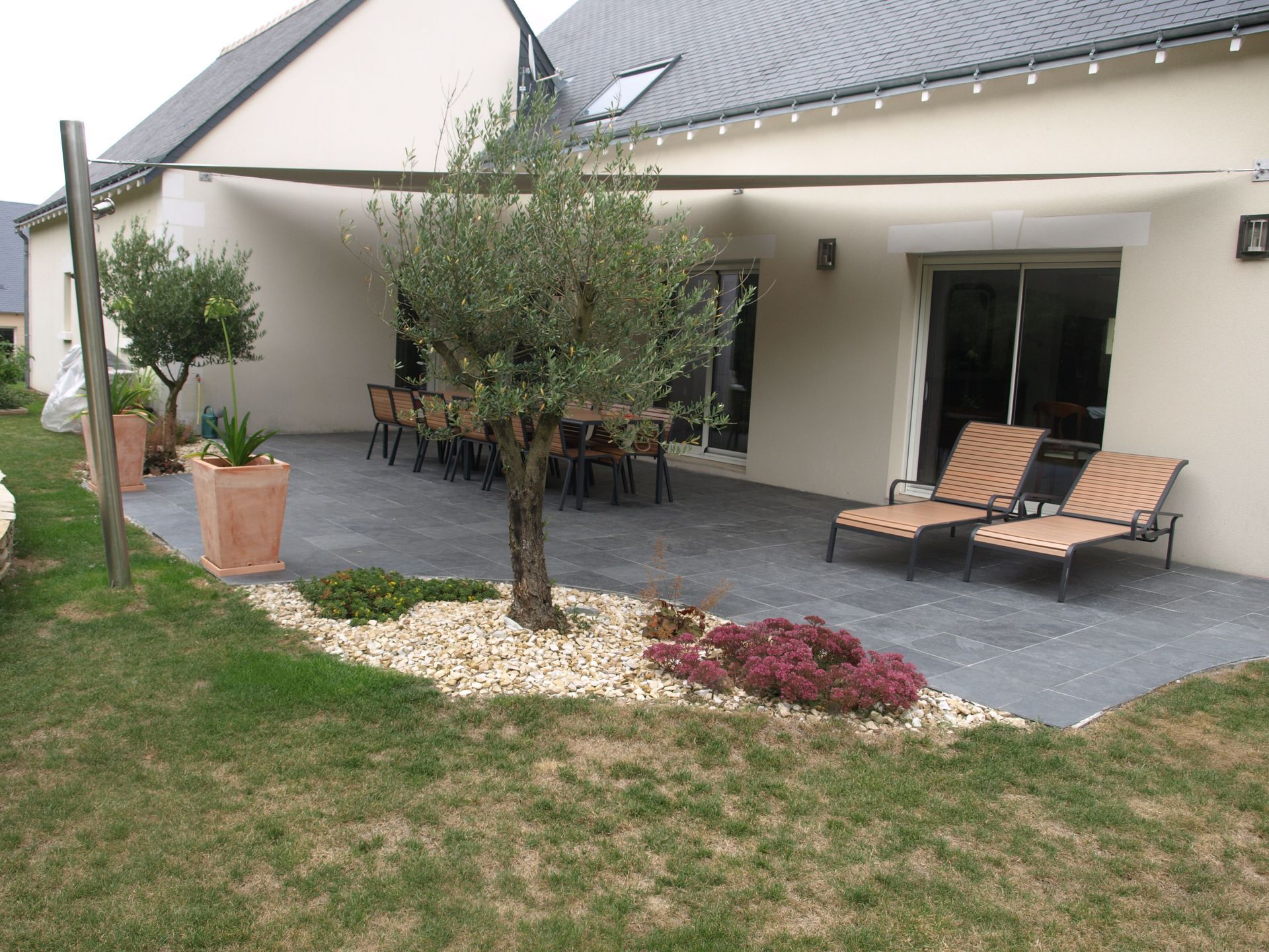 Une terrasse avec un ensemble de salle à manger et des chaises longues sous une voile d'ombrage, agrémentée d'arbres en pot et d'aménagements paysagers dans un jardin.