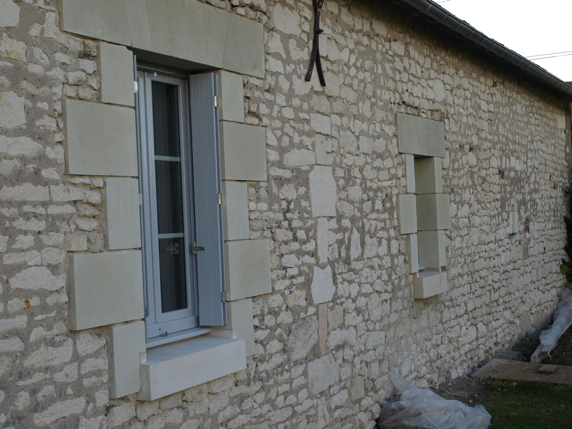 Un mur de ferme en pierre orné d'une haute fenêtre à volets bleu clair et d'une fenêtre plus petite, toutes deux encadrées de pierre lisse.
