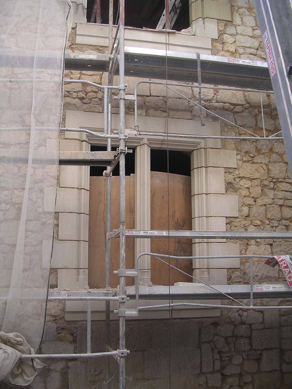 Bâtiment en pierre en cours de rénovation, avec une fenêtre condamnée encadrée de moulures en pierre ornementées, le tout entouré d'échafaudages métalliques.