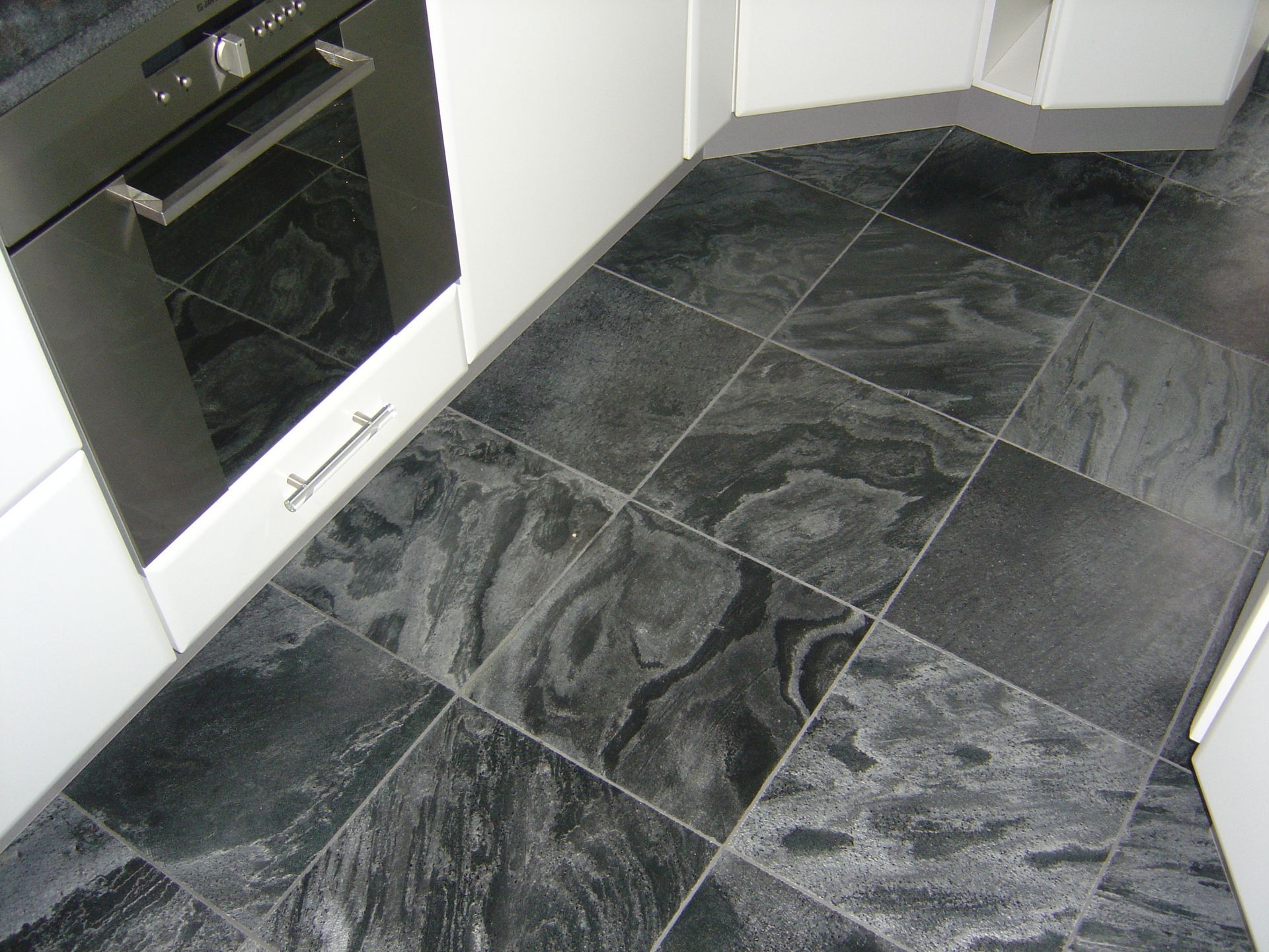 Gros plan sur des carreaux de sol de cuisine en pierre sombre à motifs, installés à côté d'un four et de meubles blancs.