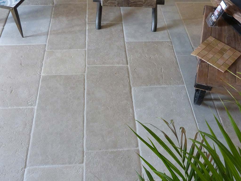 Carrelage en pierre claire disposé en motif rectangulaire, partiellement masqué par des pieds de meubles et une plante verte.