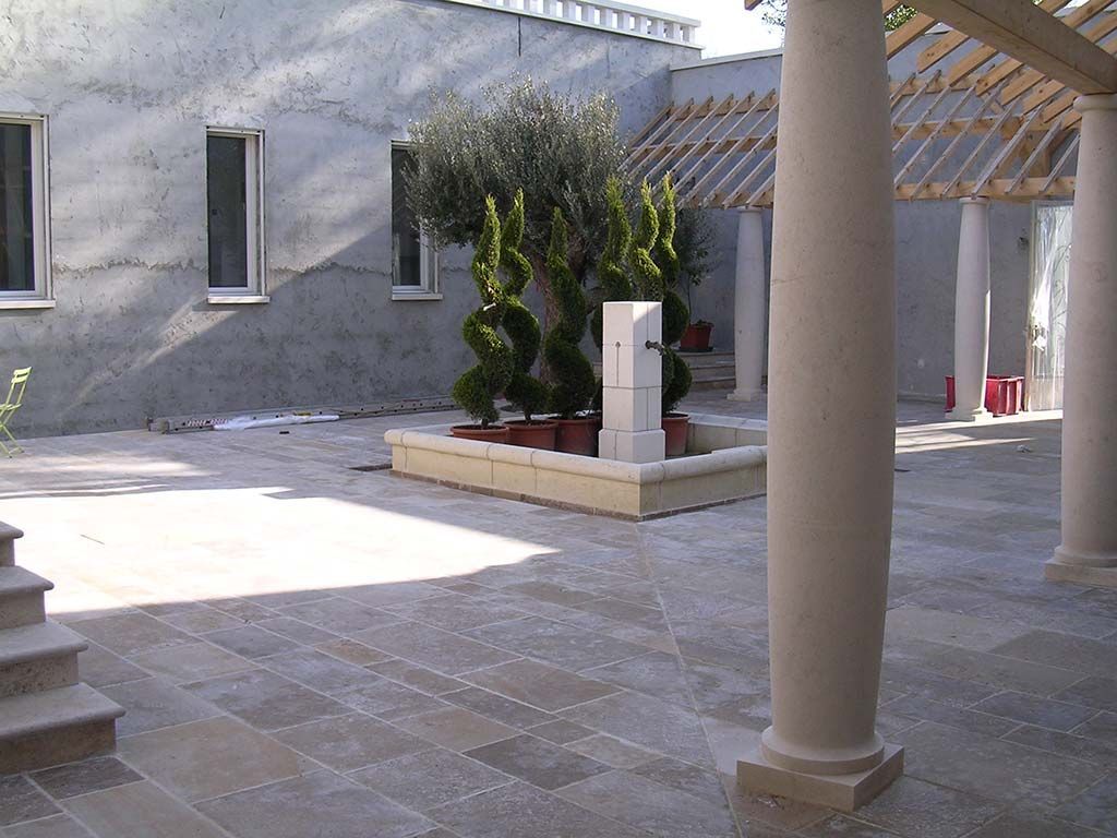 Une cour pavée et ensoleillée abrite un petit parterre de jardin avec des arbres taillés en spirale, une fontaine et des piliers de style classique.