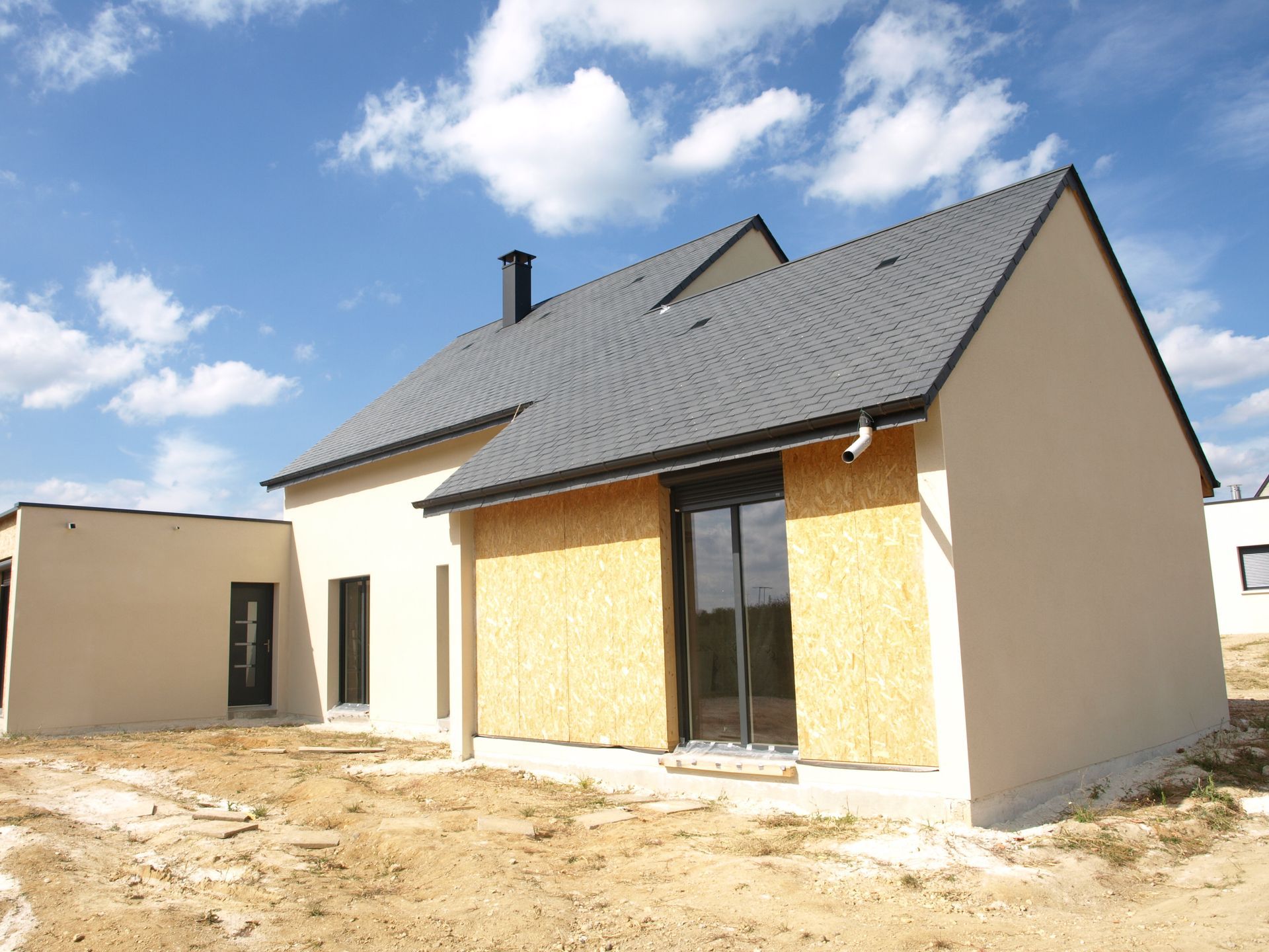 Une maison moderne avec un extérieur en stuc beige et un toit à pignons sombre, présentant une partie de mur inachevée avec des panneaux de bois.
