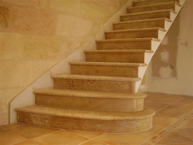 Un escalier en pierre de travertin beige avec une marche inférieure incurvée, adossé à un mur et un sol en pierre assortis.