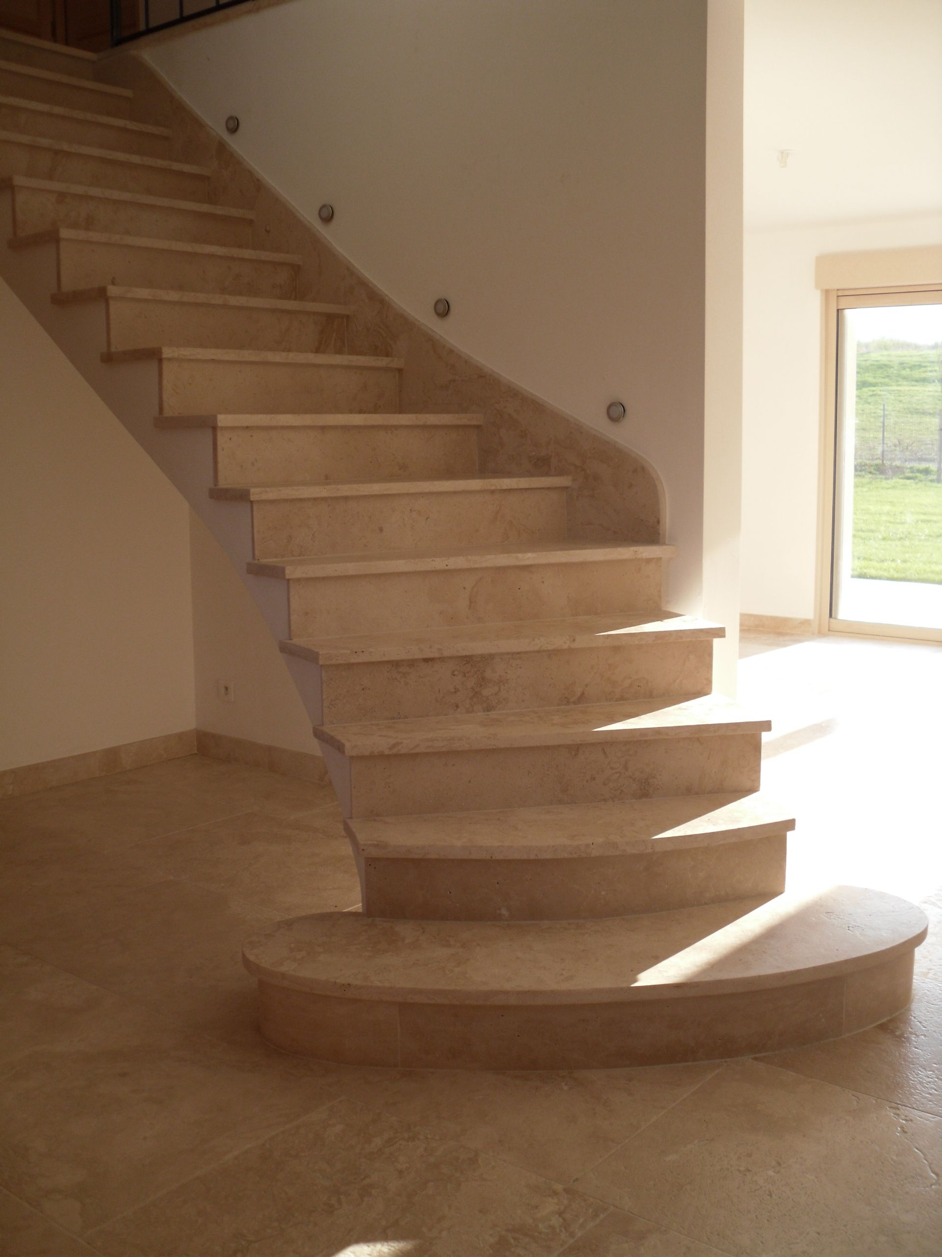 Un escalier en pierre beige, dont la première marche est incurvée, mène vers le haut, adossé à un mur clair percé de spots encastrés.