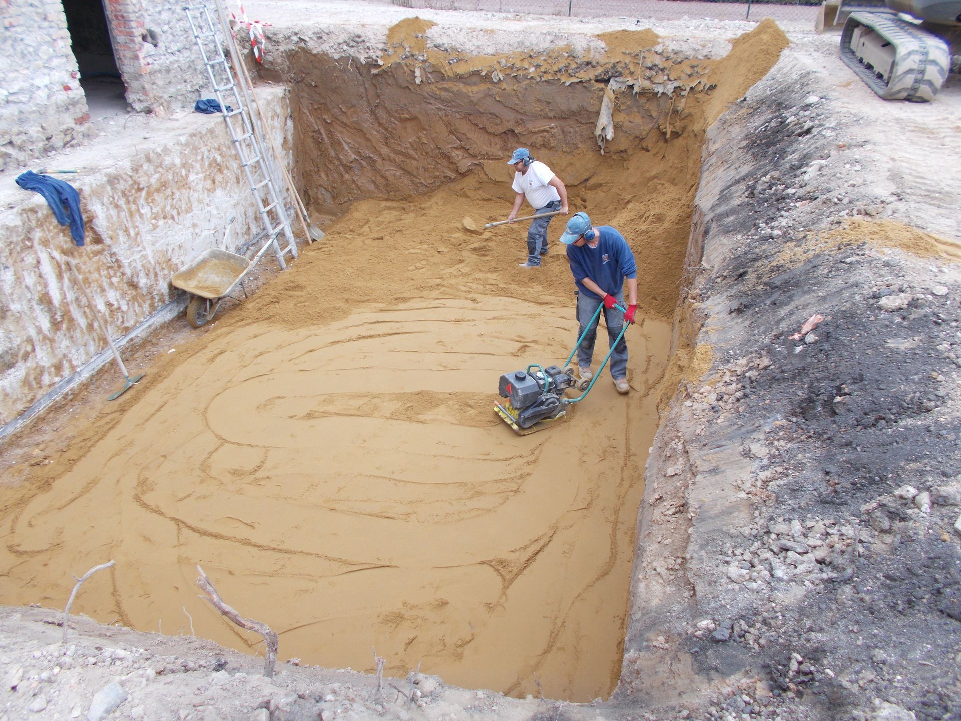 Deux ouvriers utilisent une plaque vibrante pour aplanir une base sablonneuse à l'intérieur d'une profonde fosse d'excavation pour un chantier de construction.