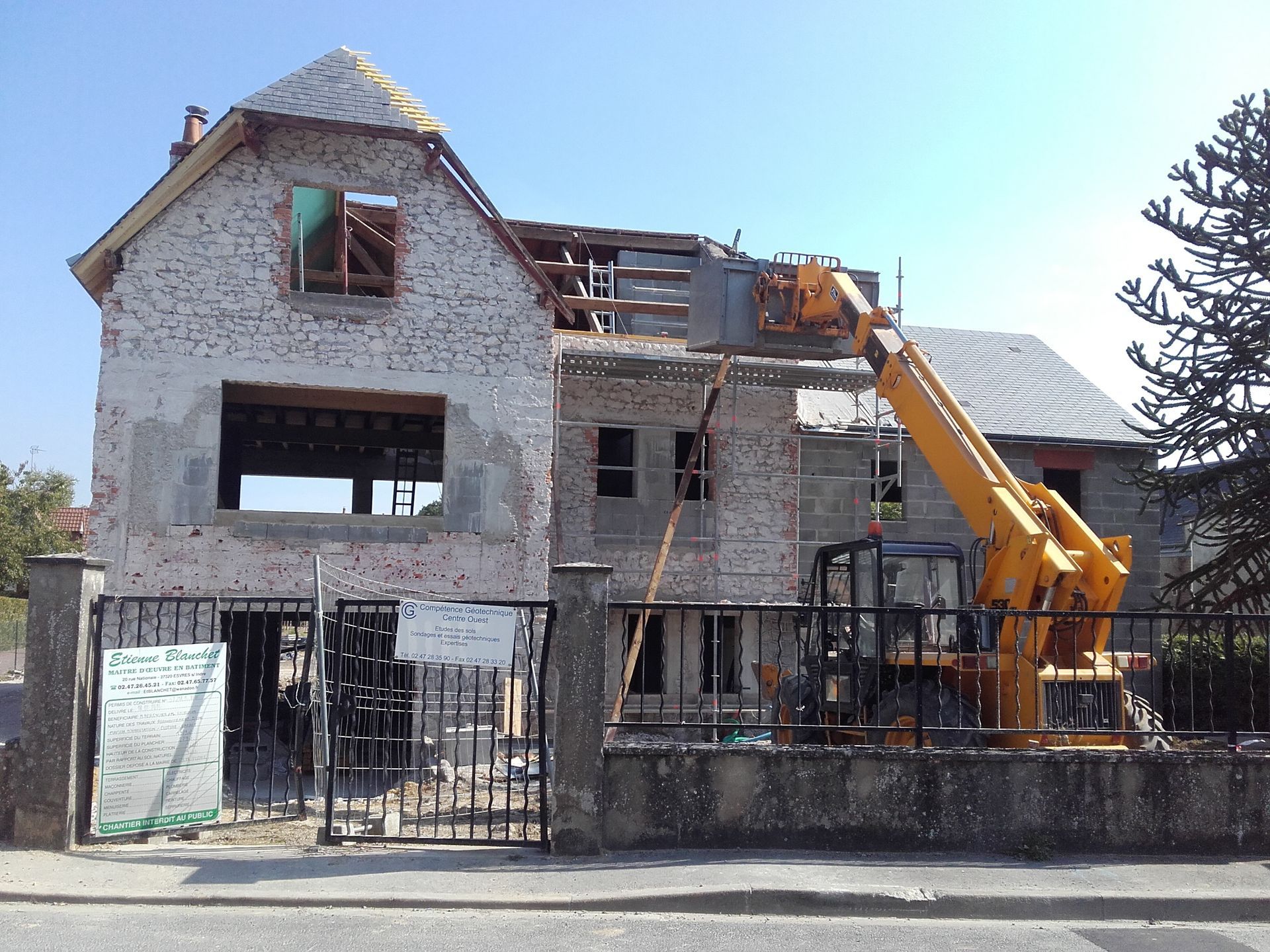 Un chariot télescopique jaune est en activité sur un chantier de construction, devant une maison partiellement démolie dont la façade en briques est apparente.